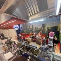 View from behind the counter: crepe griddles and prep station, with the painted skyline mural and guests seated beyond