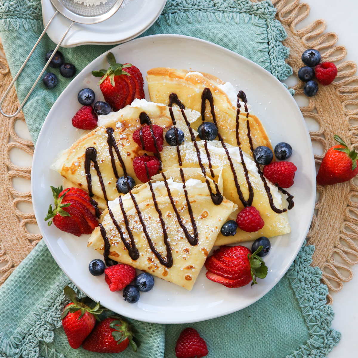 A plate of folded crêpes dusted with icing sugar and drizzled with chocolate, ringed with strawberries, raspberries and blueberries